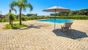 Zona de piscina con patio de ladrillo, palmeras, una silla de descanso y una sombrilla.
