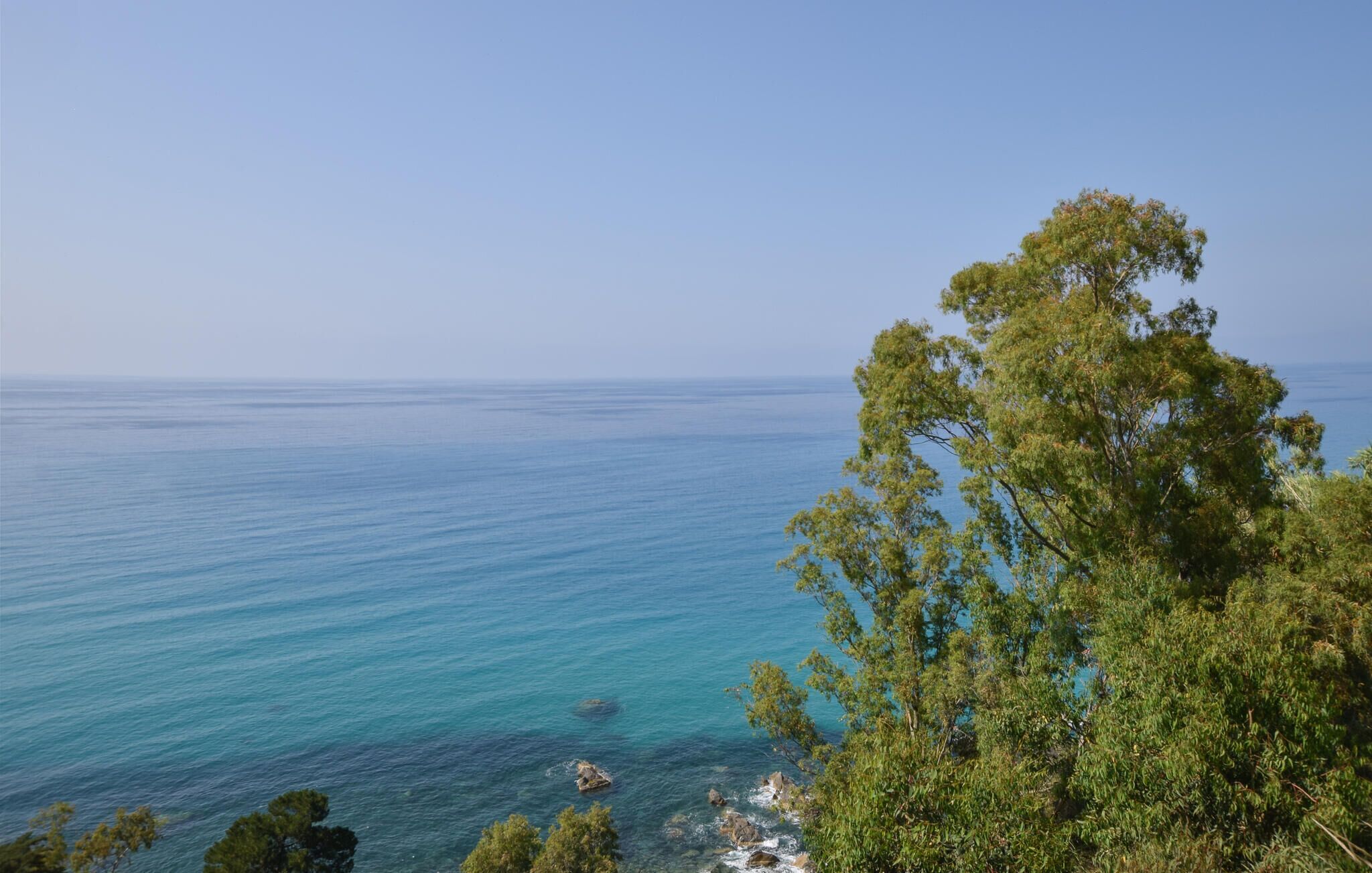 A coastal landscape with clear blue sea, rocky outcrops, and dense green foliage.