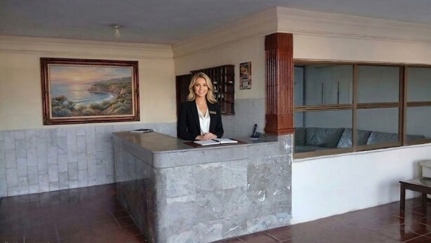 Un mostrador de recepción con encimera de mármol, un cuadro con un paisaje colgado encima, y una ventana con vista a un edificio.