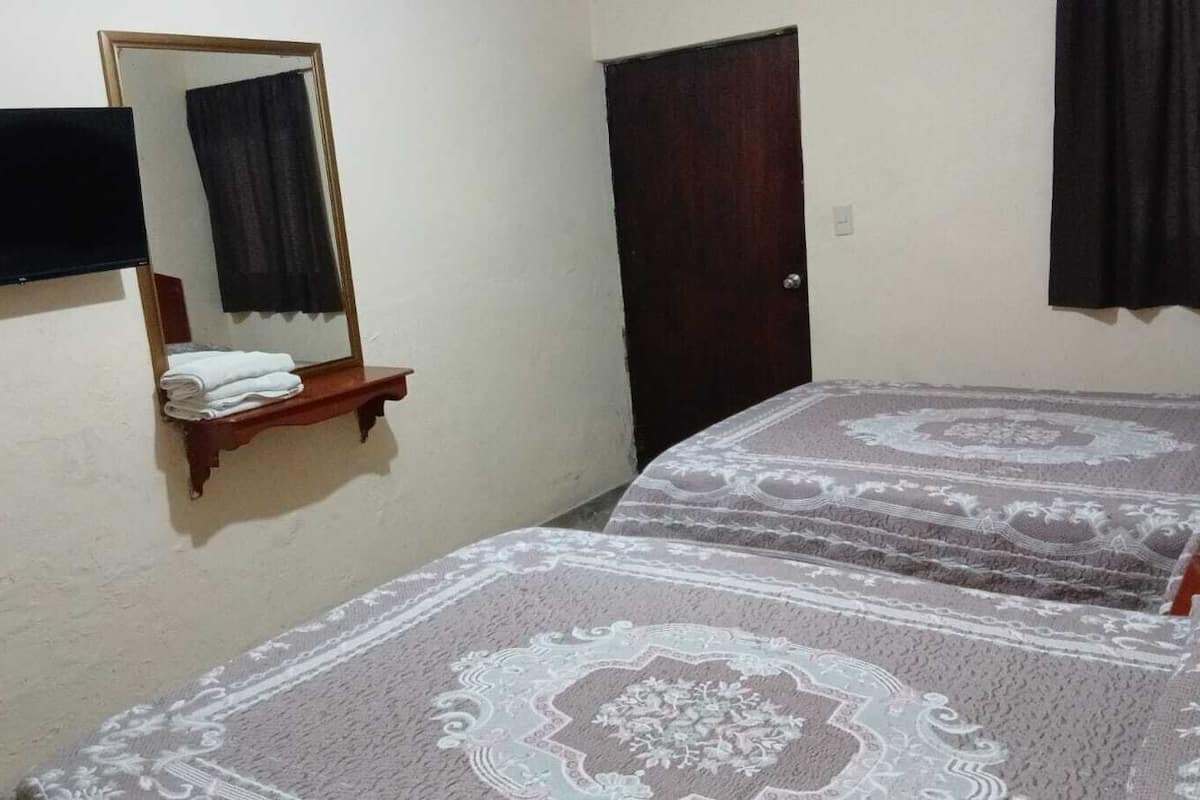 A bedroom with two beds, a wooden shelf with folded towels, a dark door, and a mounted TV.