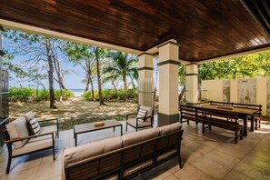 An outdoor patio with a wooden ceiling, a dining table, and chairs, overlooking a beach with palm trees and the ocean. - Casa Palmas. Beachfront Villa Escape w/ Private Pool & Direct Beach Access (Tamarindo)