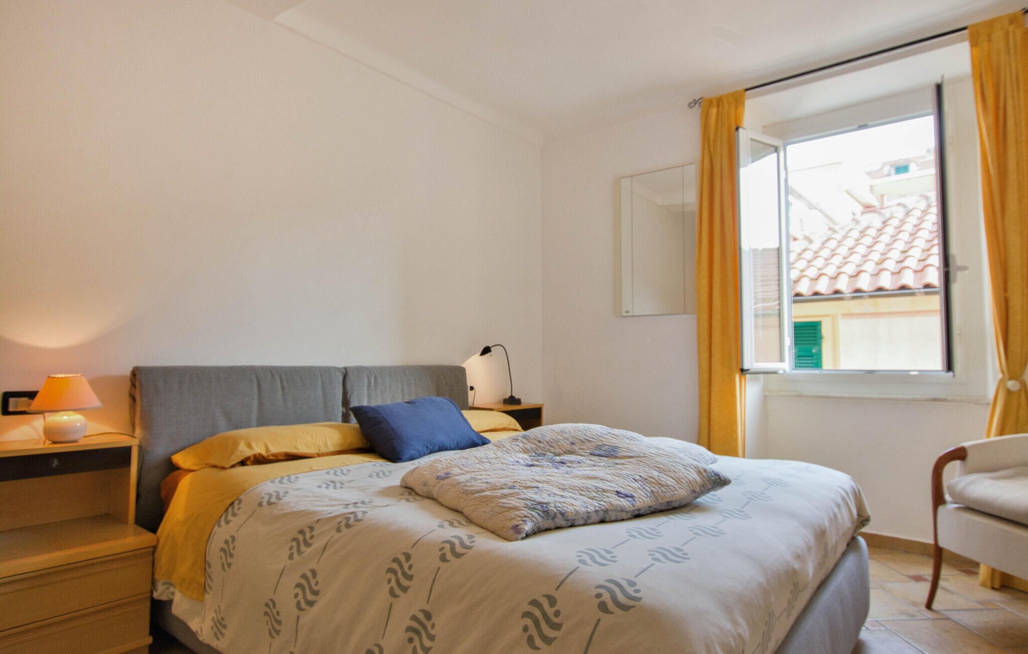 Un dormitorio con una cama, una mesita de noche con lámpara, una silla y un ventanal con cortinas.