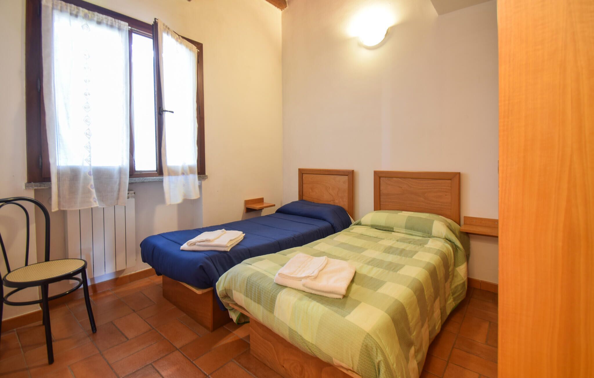 A room with two beds, a chair, a window with curtains, and a wall-mounted light fixture.