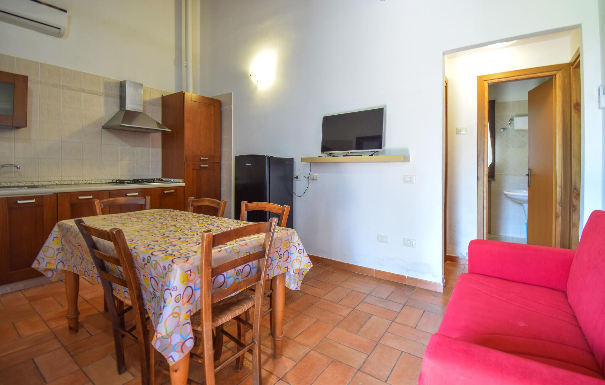 A compact kitchen with a dining table, chairs, a red couch, and a television mounted on the wall.