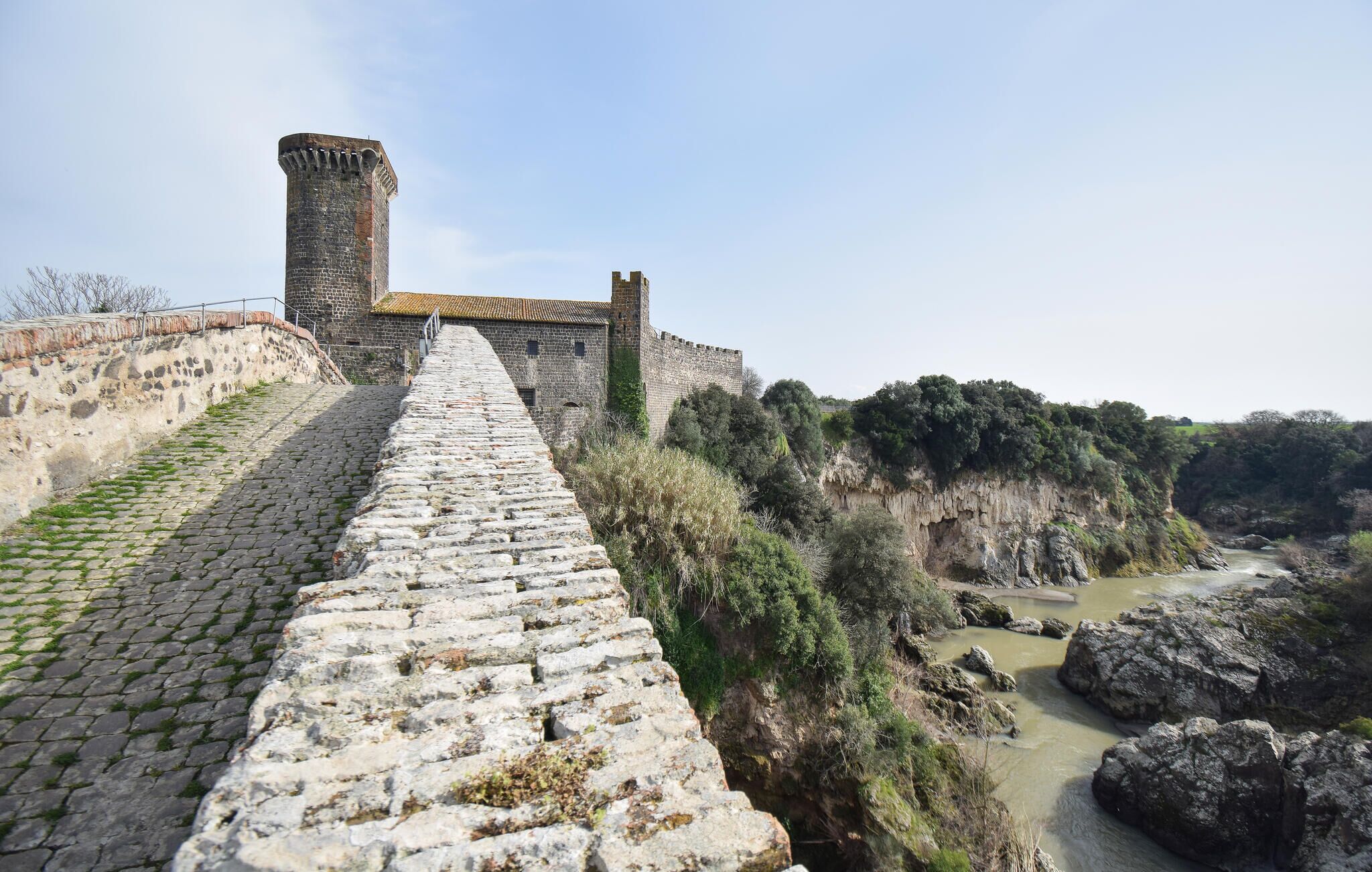 A stone pathway leading to a historic stone building with a tower, surrounded by natural landscape and a river.