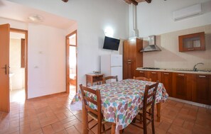 Eine Küche mit Holdelementen, einer gefliesten Spritzschutzwand, einem Herd und einem Spülbecken. Es gibt einen Esstisch mit Stühlen und einen an der Wand befestigten Fernseher.