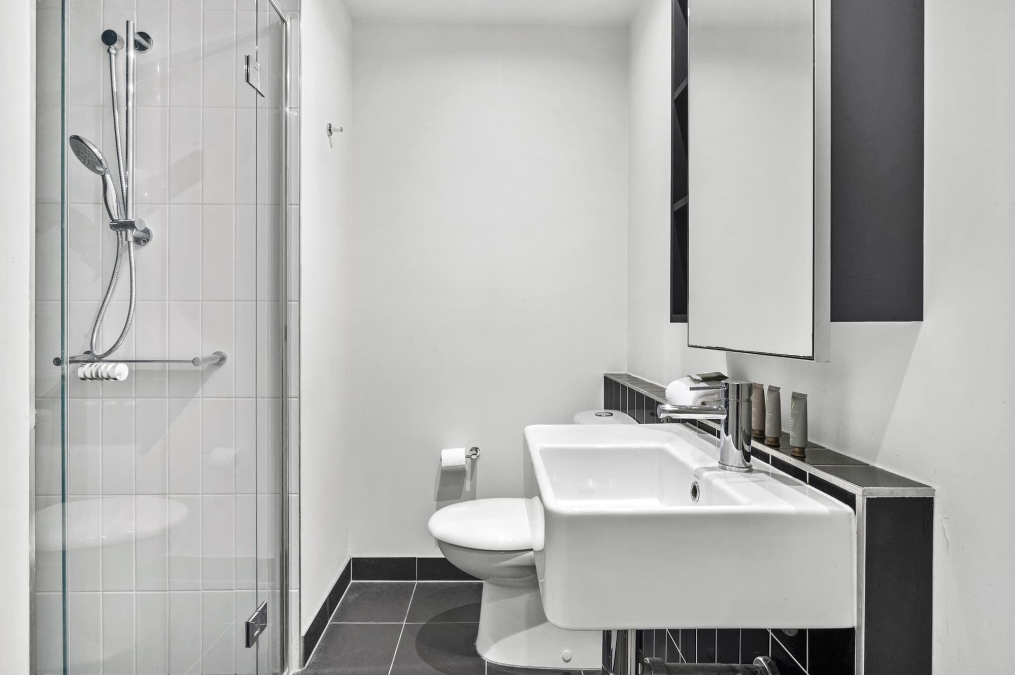 A modern bathroom with a glass shower enclosure, a white toilet, a sink with a mirror above, and a black tiled floor.