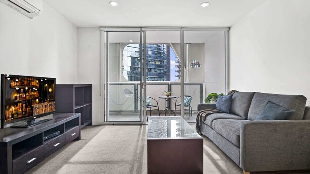 Ein modernes Wohnzimmer mit Flachbildfernseher, grauem Sofa, Glastisch und einem Balkon mit Blick auf die Stadt.