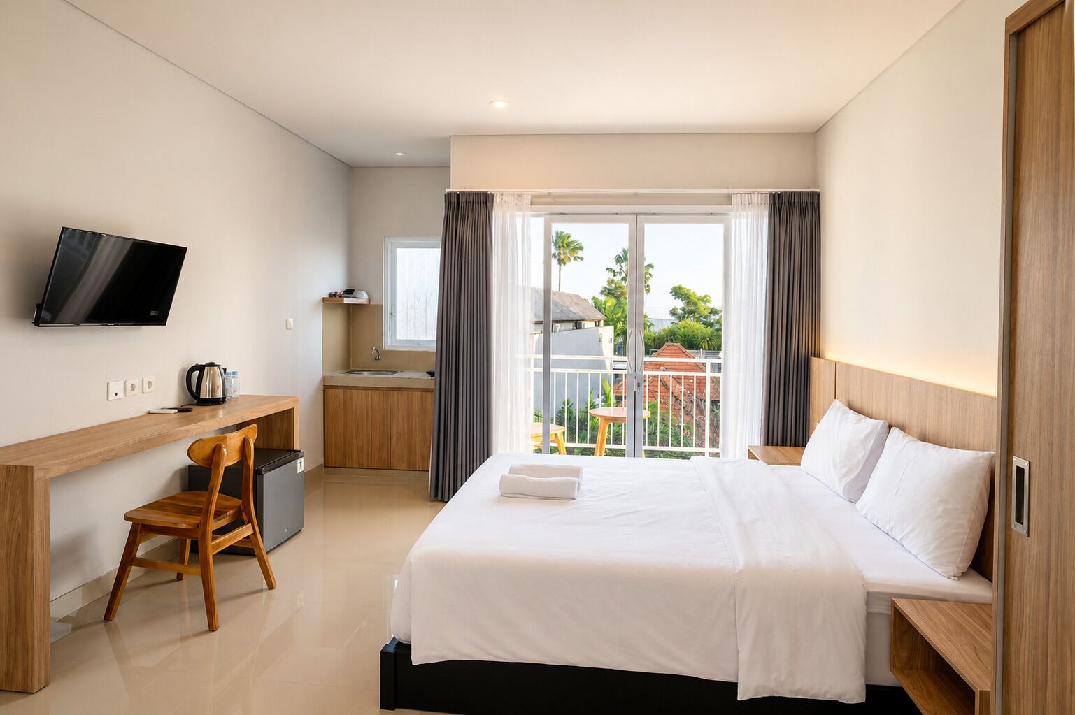 Habitación de hotel con cama, escritorio, silla, televisor, kitchenette y balcón con vistas.