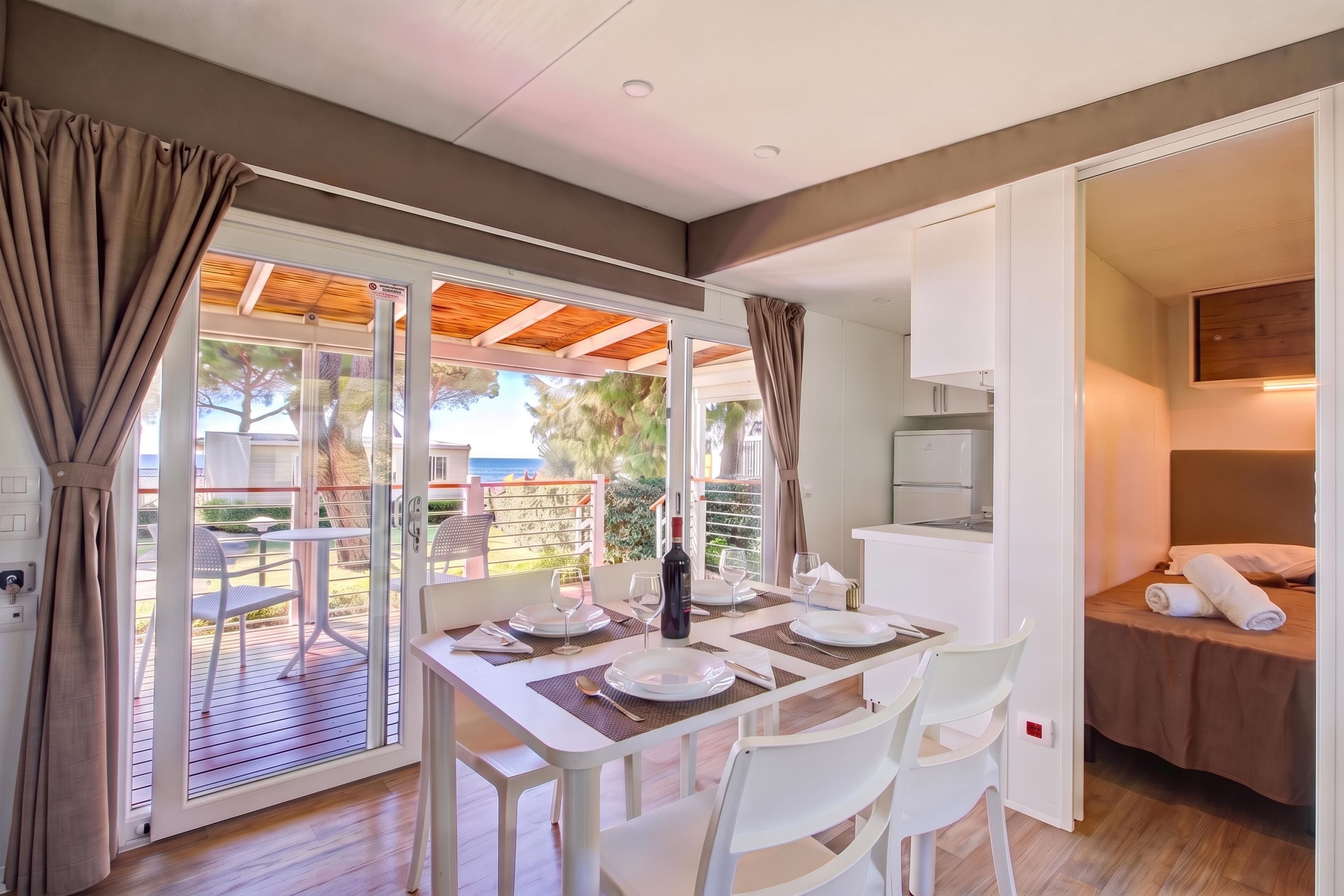 A dining area with a table set for two, a bed with a white headboard, and a view of a garden through a sliding glass door.