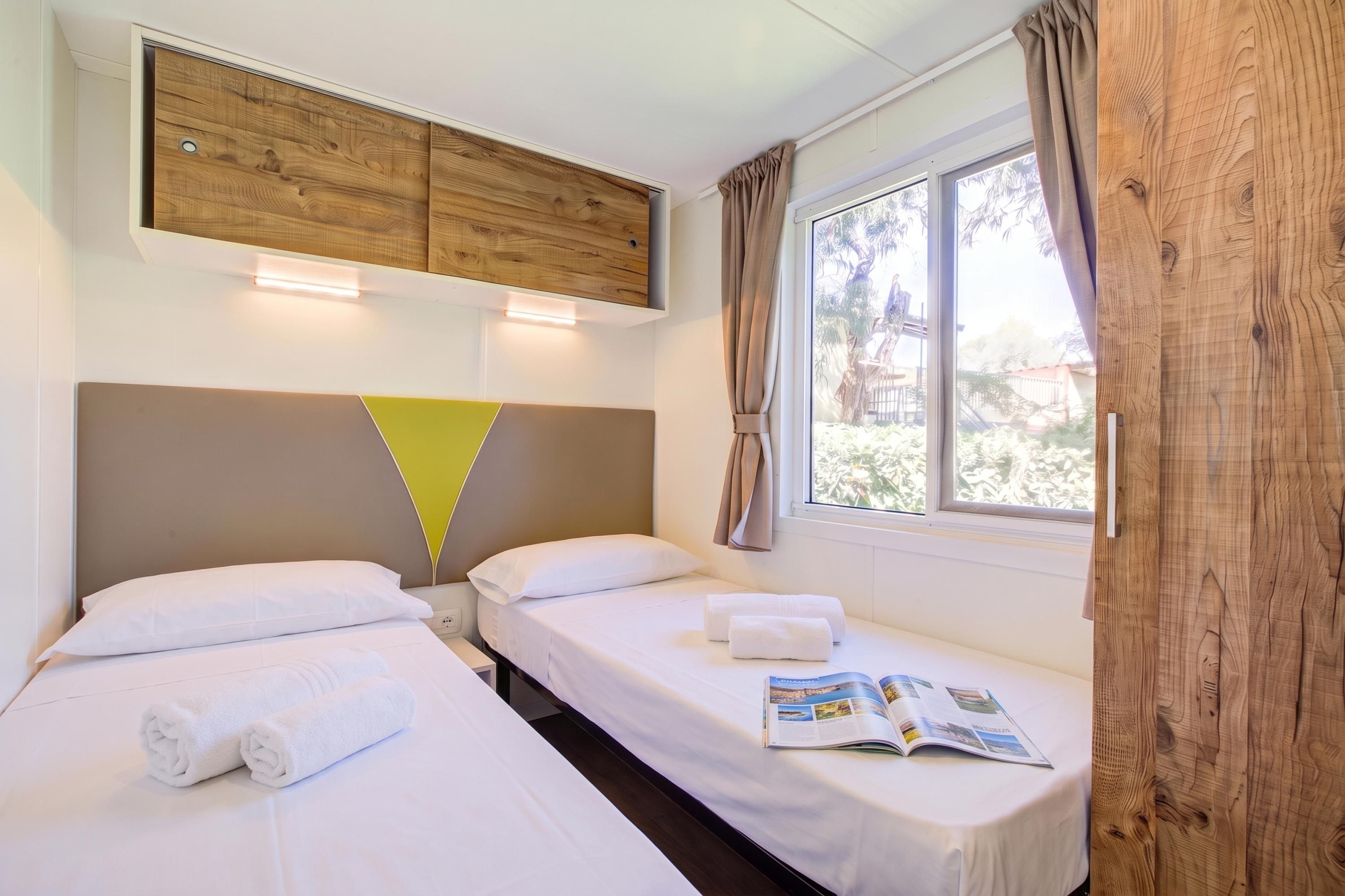 Habitació d'hotel amb dos llits, un capçal de llit de fusta, una finestra amb cortines i una revista damunt del llit.