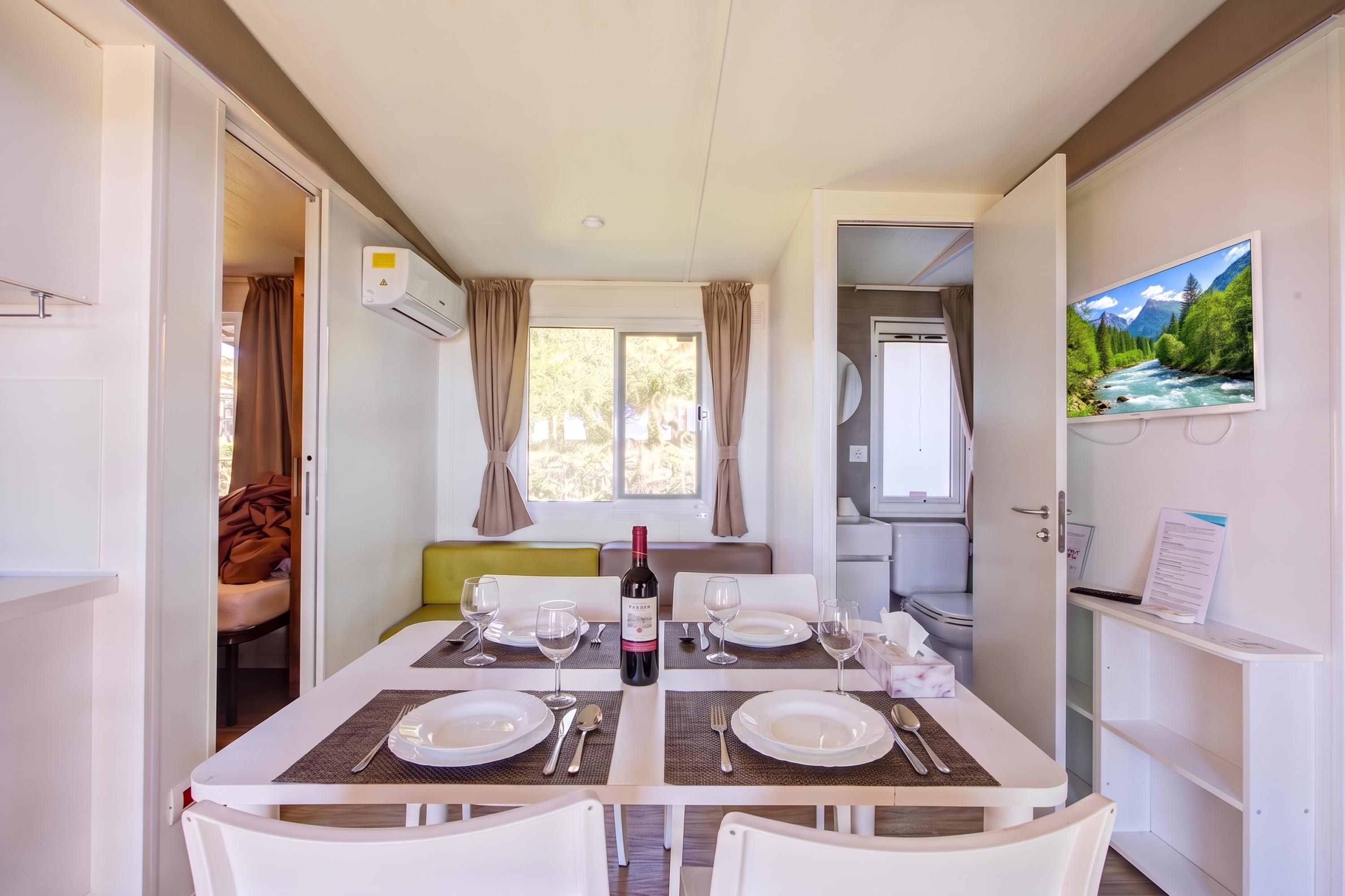 Un comedor con una mesa dispuesta para dos personas, una botella de vino y un televisor que muestra un paisaje panorámico.