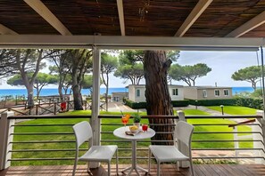 Un balcón con dos sillas blancas, una mesa con bebidas y fruta, y vistas al mar y a los árboles.