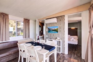 Habitació amb taula de dinar per a dues persones, sofà, televisió penjada a la paret i llit amb vista a l'exterior.