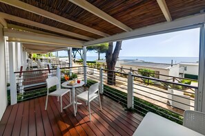 A covered outdoor area with a wooden deck, white chairs, a table, and a view of the sea.