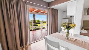 Ein modernes Zimmer mit Holzboden, weißer Einrichtung, einem Esstisch mit Stühlen, einer Küchenzeile und einem Balkon mit Blick ins Grüne.