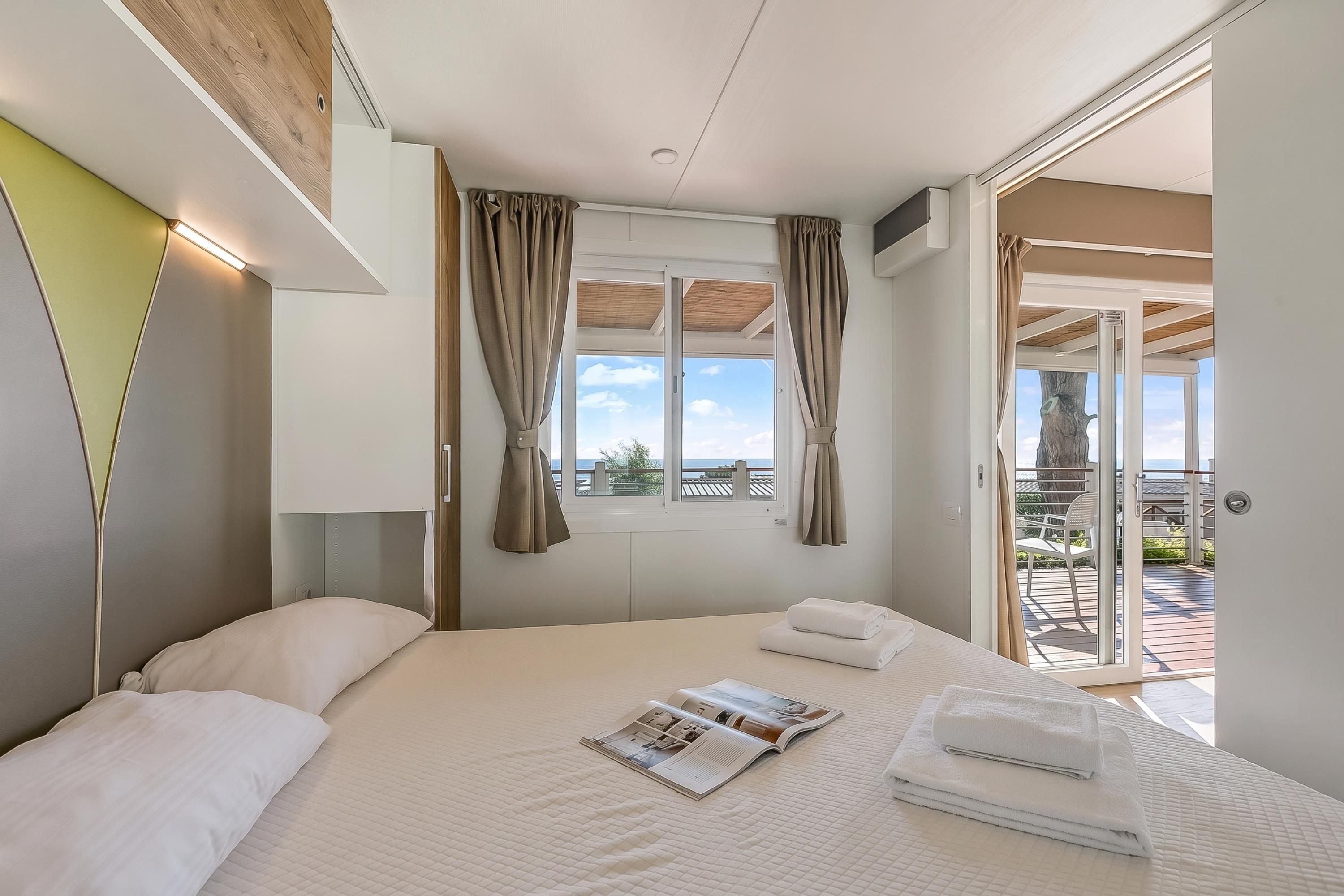 Een hotelkamer met een bed, een raam met gordijnen, een deur die naar een buitengebied leidt en een tijdschrift op het bed.