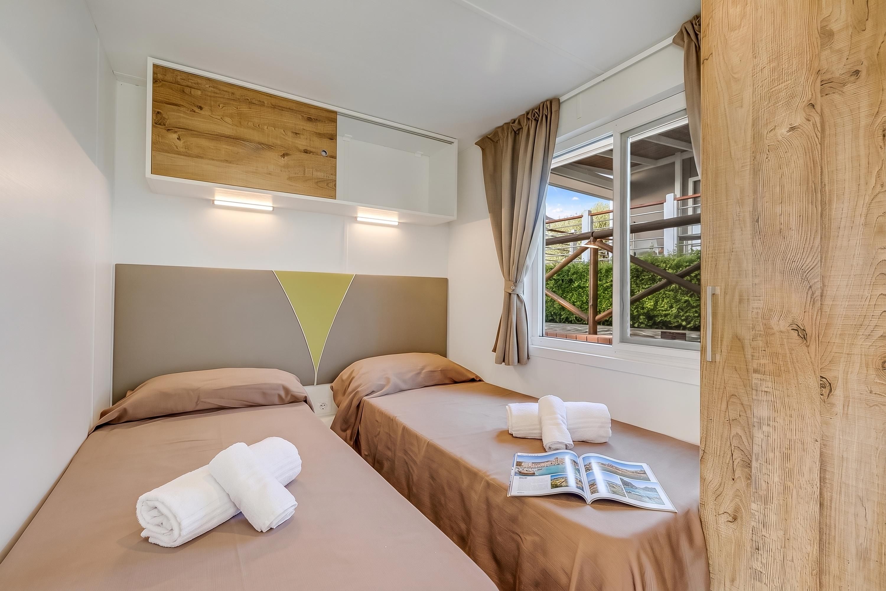 Une chambre d’hôtel avec deux lits, une fenêtre avec des rideaux, une tête de lit en bois et un magazine posé sur le rebord de la fenêtre.