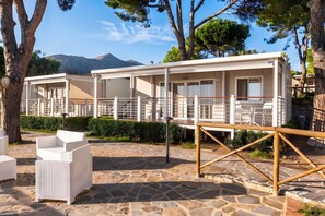 A row of white cabins with balconies, set on a stone-paved area with trees and a mountain in the background.
