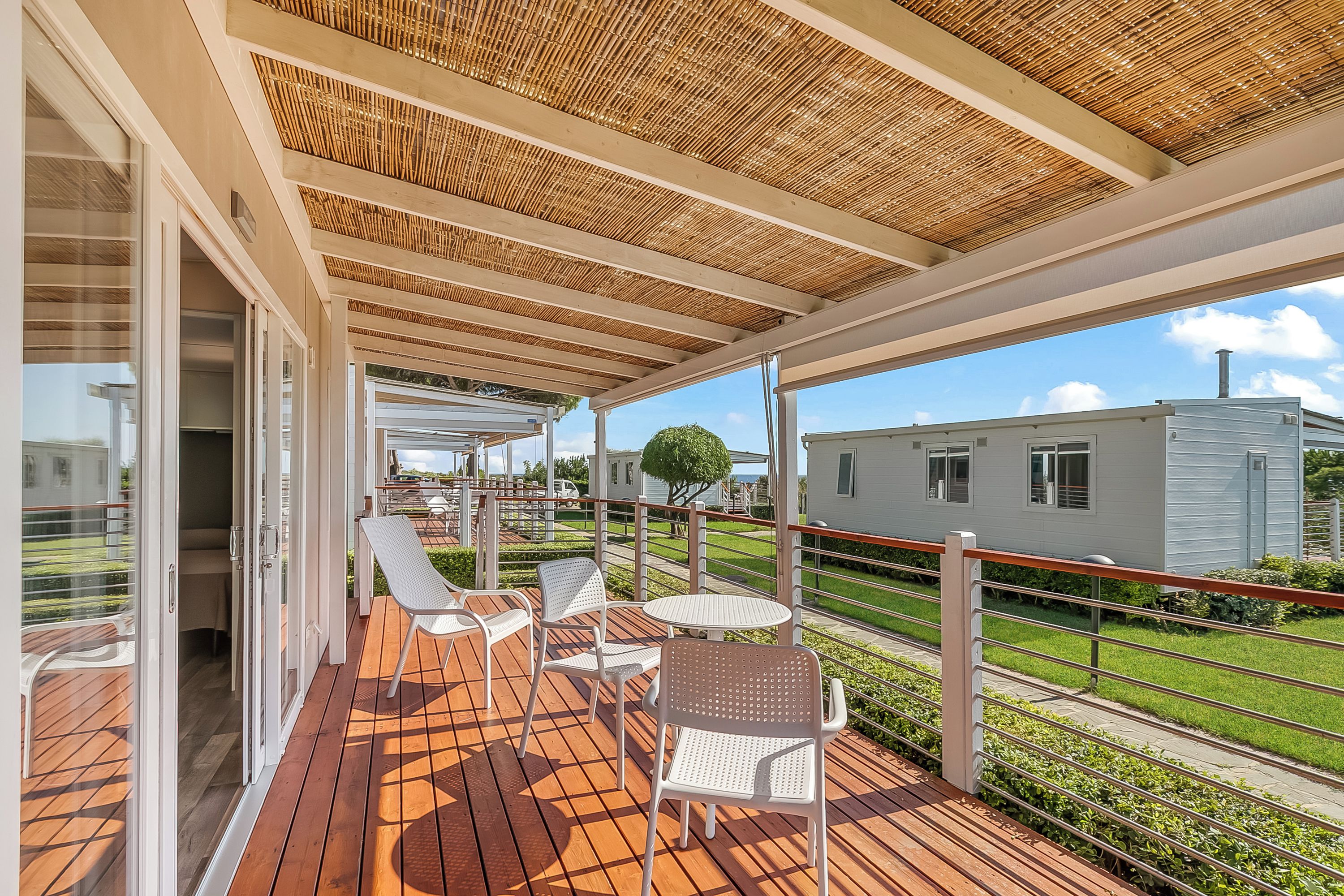 Un espacio al aire libre techado con una terraza de madera, sillas de metal blanco y una mesa.