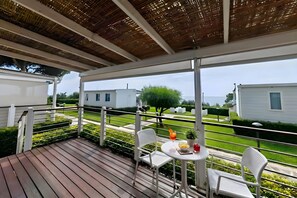 Un espacio exterior cubierto con una terraza de madera, sillas de metal blanco, una pequeña mesa redonda y una planta en maceta.