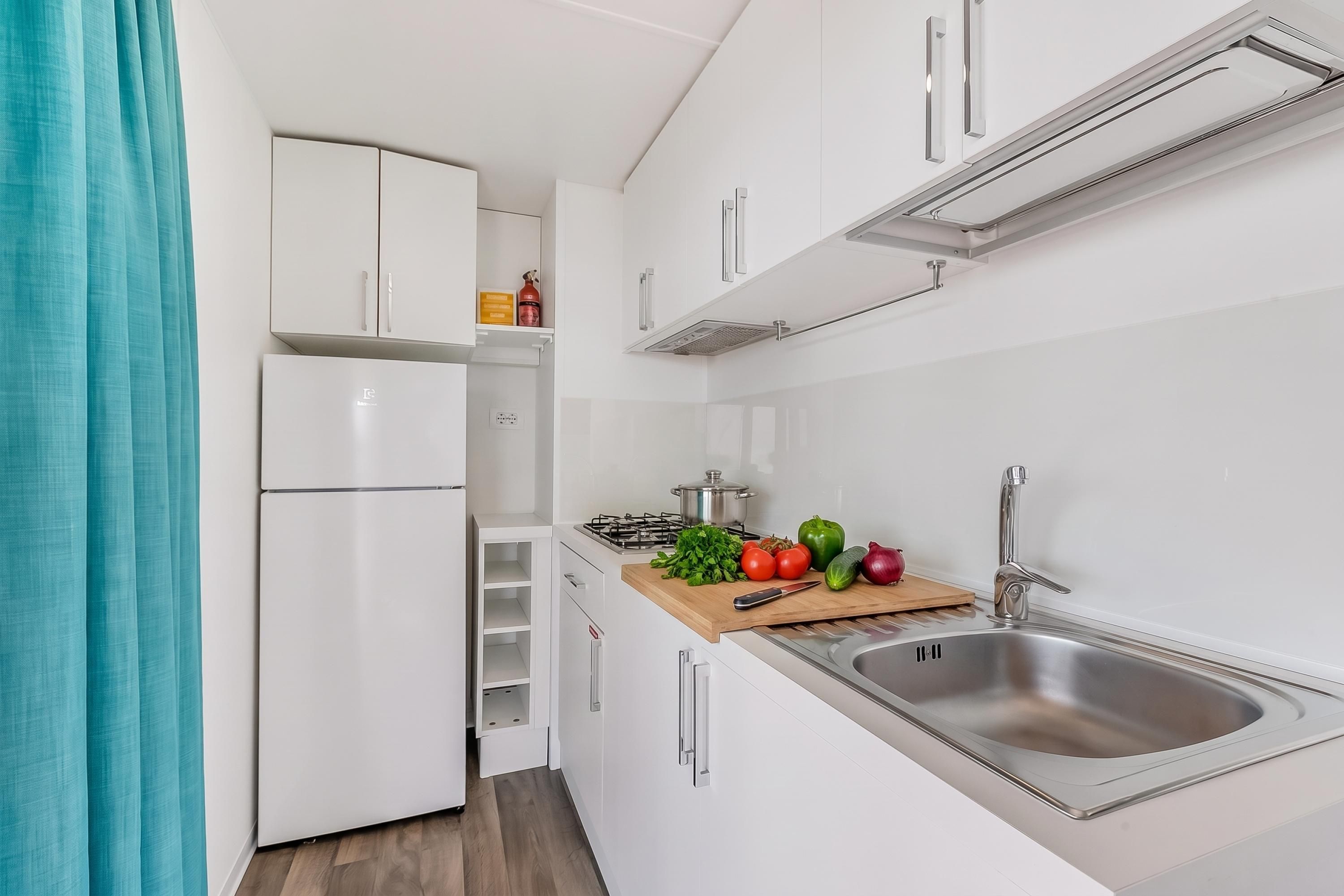 Une cuisine compacte avec des meubles blancs, un évier en acier inoxydable, une cuisinière à gaz et un plan de travail en bois sur lequel sont disposés des légumes frais.