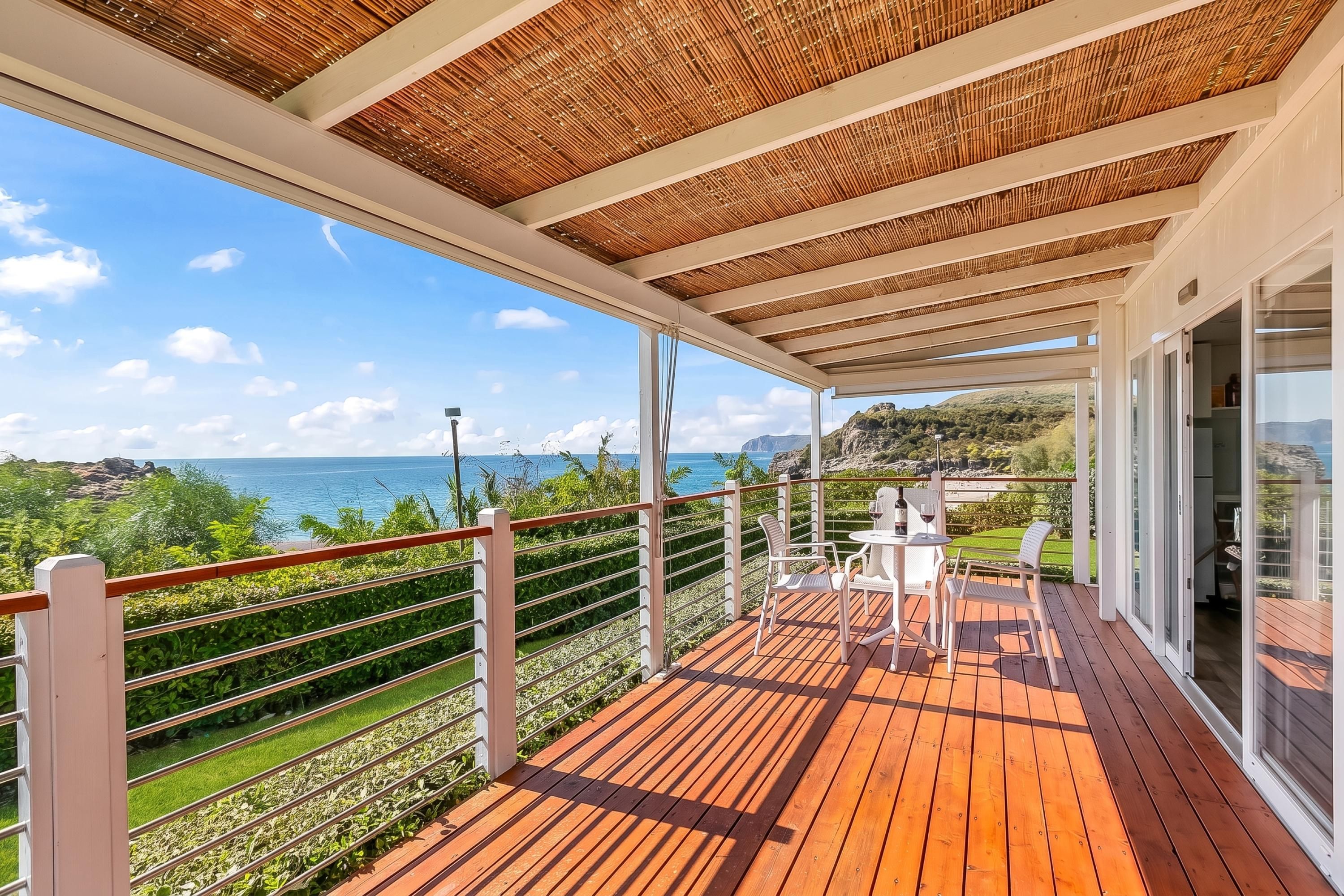 A covered outdoor area with a wooden deck, white chairs, and a table, overlooking a coastal landscape.