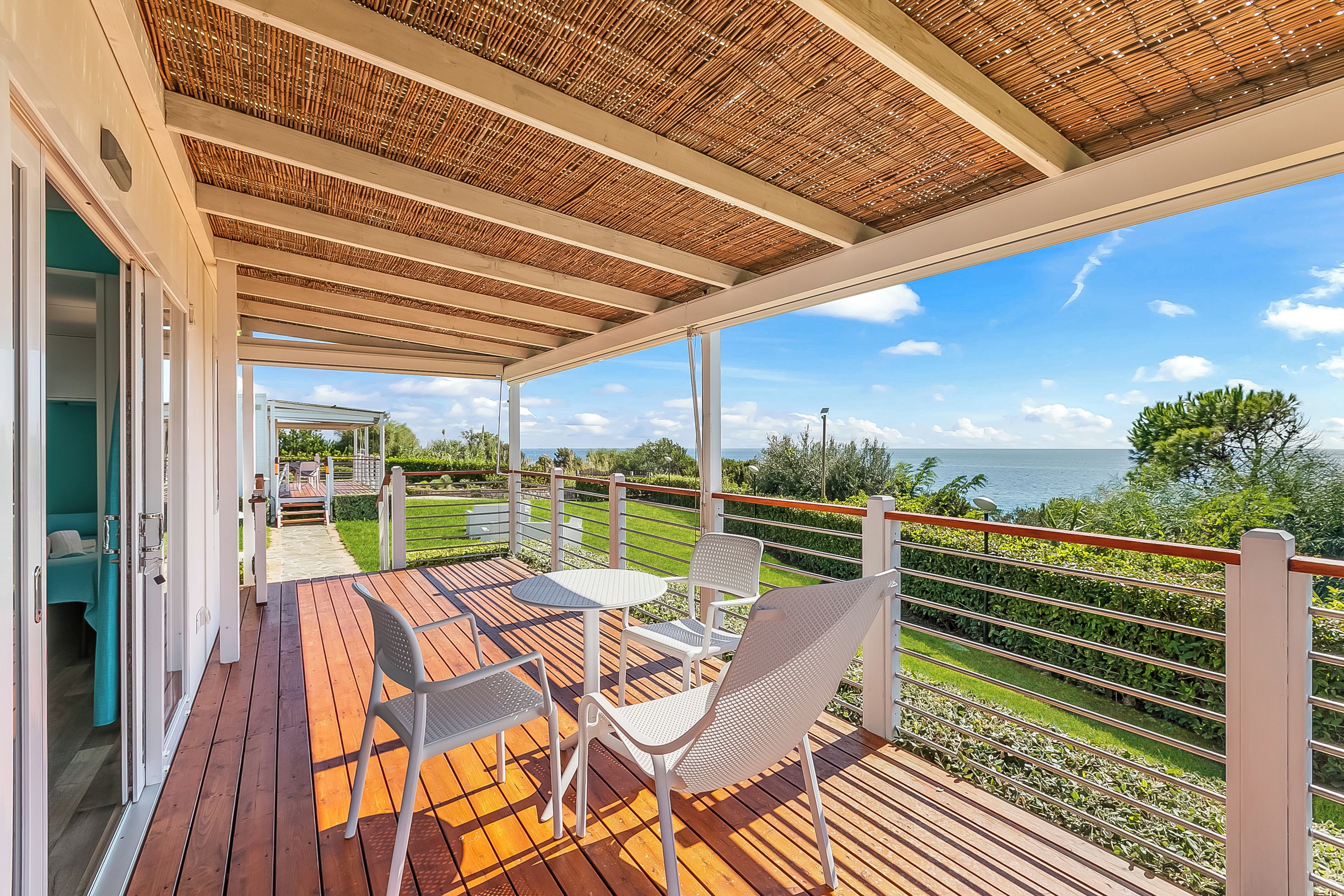 Un espacio exterior cubierto con una terraza de madera, sillas de metal blanco y vista al mar.