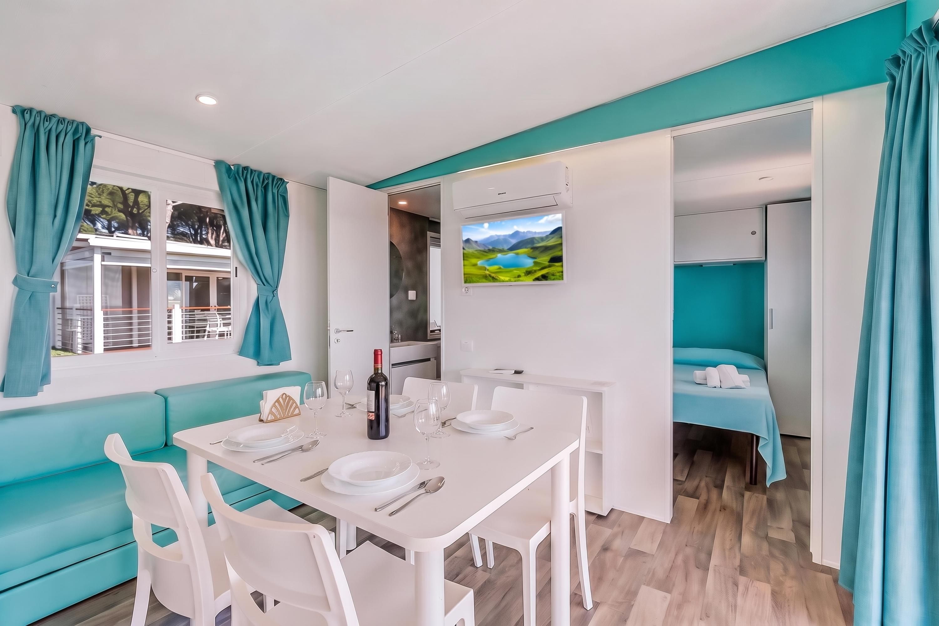 A modern, well-lit room with a dining table set for two, a sofa, and a TV mounted on the wall displaying a scenic landscape.