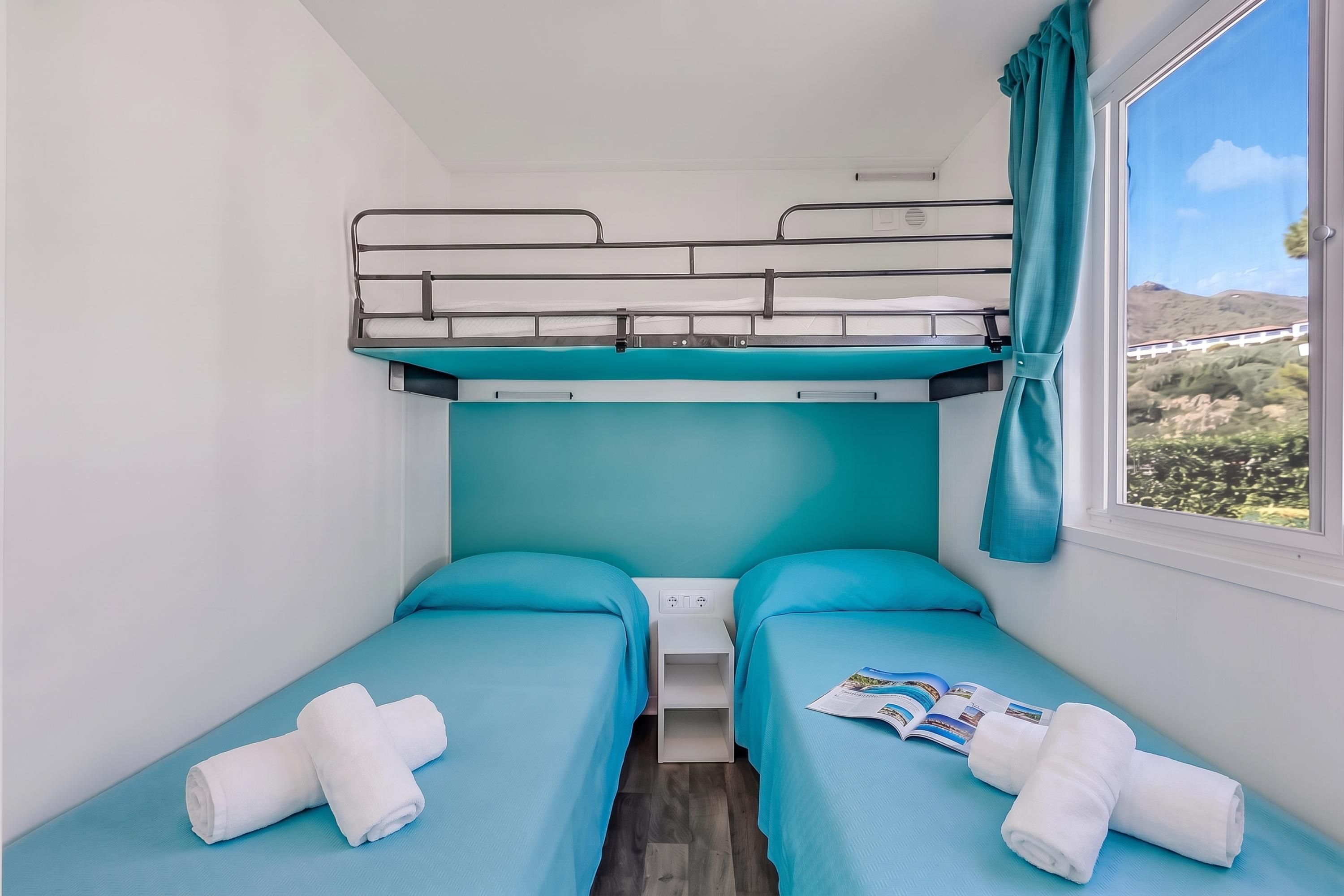 A compact room with two single beds, a bunk bed above, a window with a view of a mountainous landscape, and a small bedside table with a magazine.
