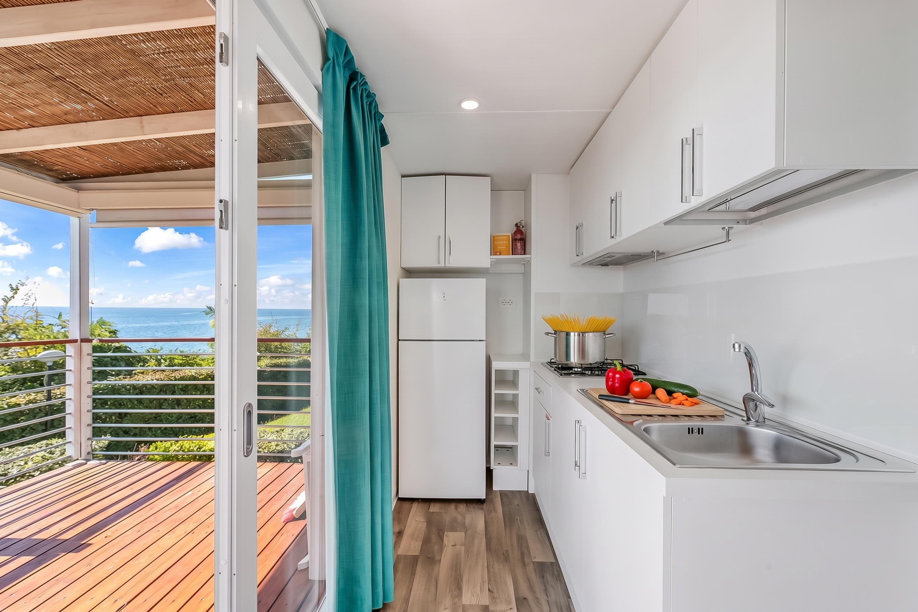 Une cuisine compacte avec vue sur la mer, un balcon doté d’un plancher en bois et un rideau de couleur turquoise.