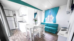 Eine kompakte Küche mit weißen Schränken, einem Esstisch mit Stühlen, einem blaugrünen Sofa und einem Fenster mit blauen Vorhängen.