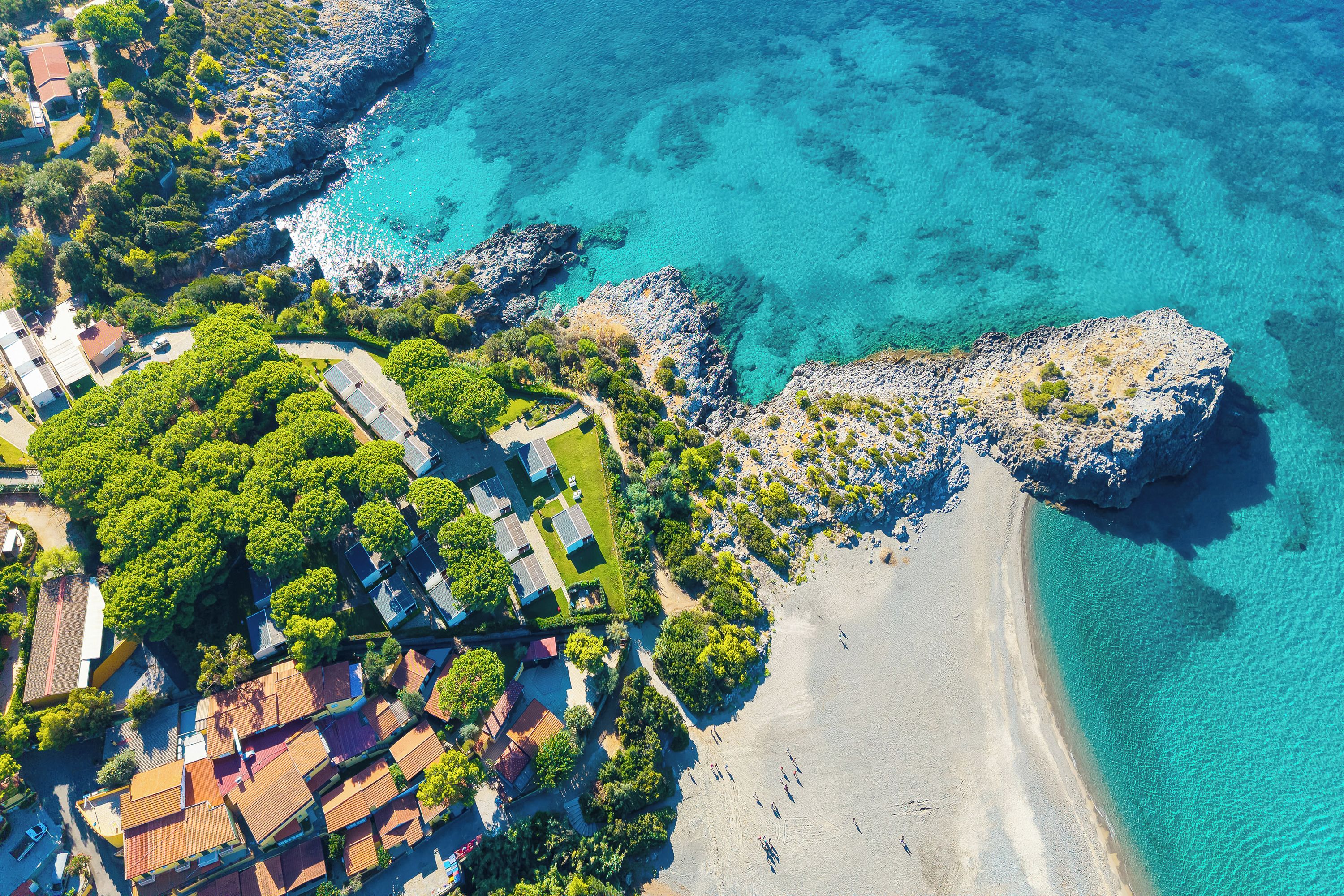 Vue aérienne d’une zone côtière avec des bâtiments, une plage et une eau d’un bleu limpide.
