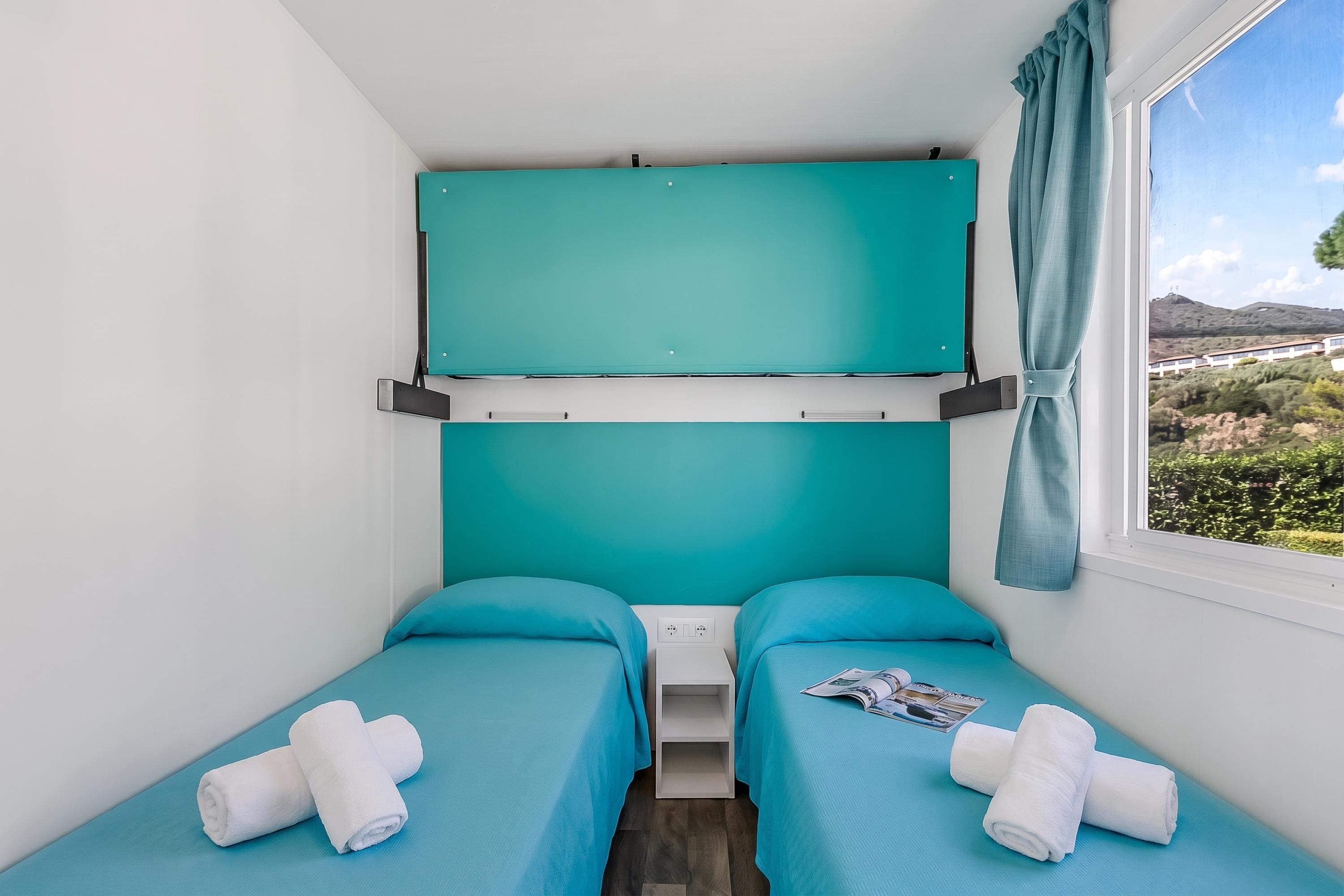 A small, single-room hotel room with two beds, a window with a view of a landscape, and a teal-colored headboard.