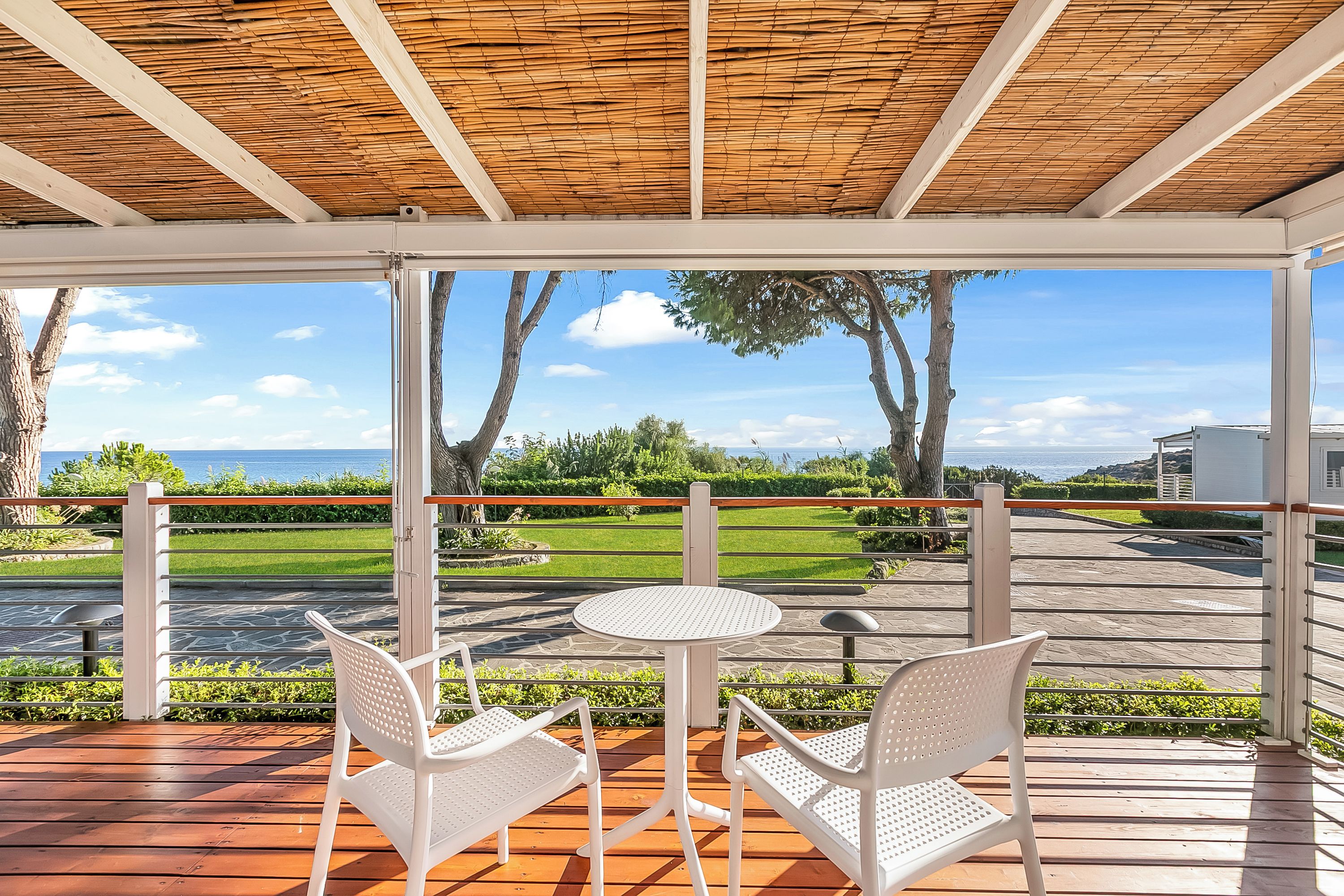 Un espace extérieur aménagé, abrité et doté de deux chaises blanches et d’une table, offrant une vue sur un jardin paysager et la mer.
