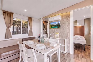 Un comedor moderno con una mesa lista para dos personas, un televisor montado en la pared, y una cama con cabecera de madera en la habitación contigua.