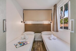 Een kleine kamer met twee eenpersoonsbedden, een raam met gordijnen, een plafondlamp en een tijdschrift op de vloer.