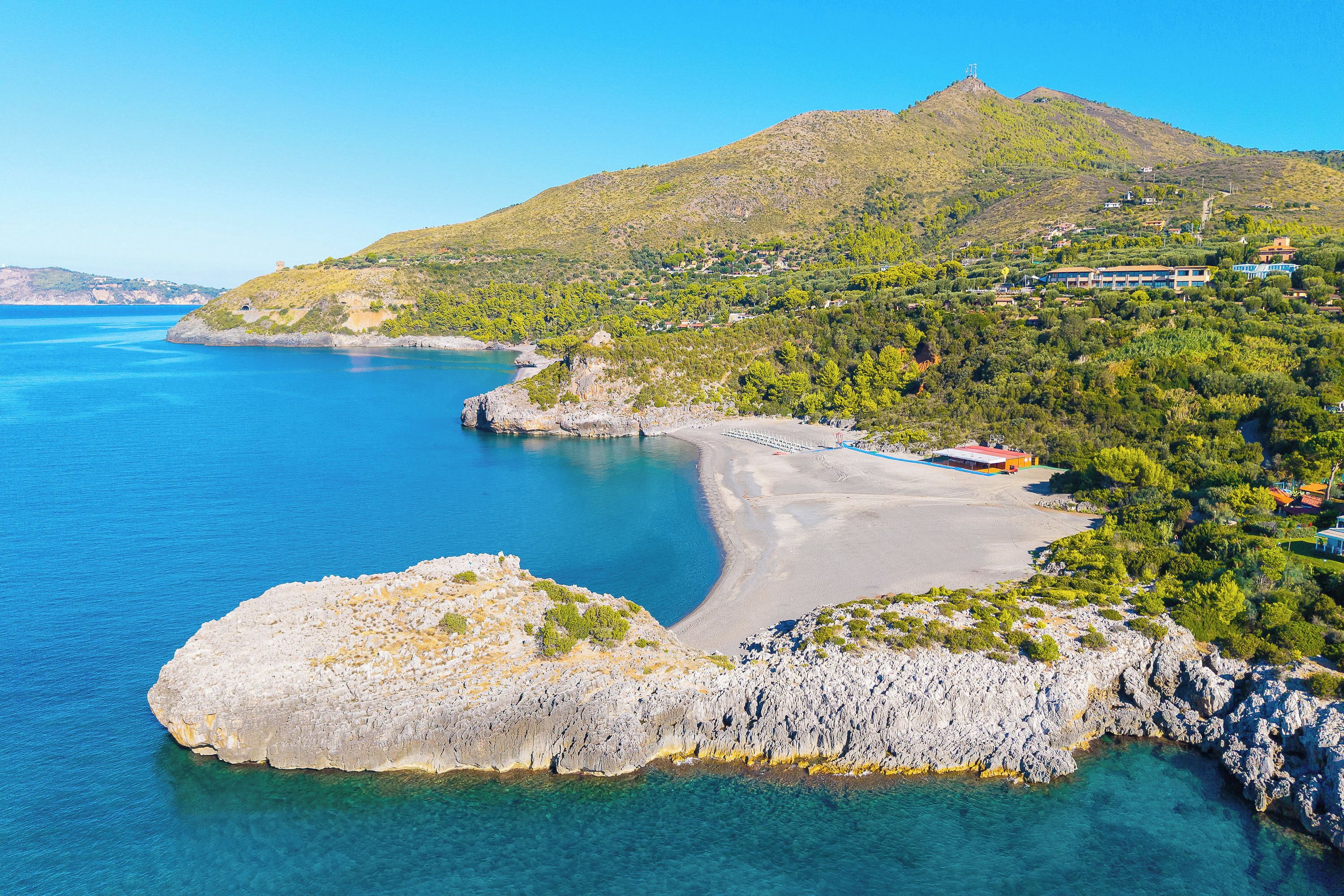 Un paisaje costero con mar azul transparente, una costa rocosa y una colina con edificios dispersos.
