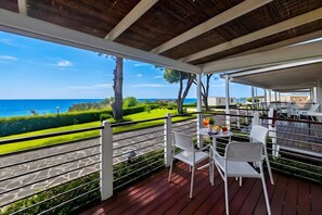 A covered outdoor seating area with a view of the sea, wooden flooring, and white chairs.