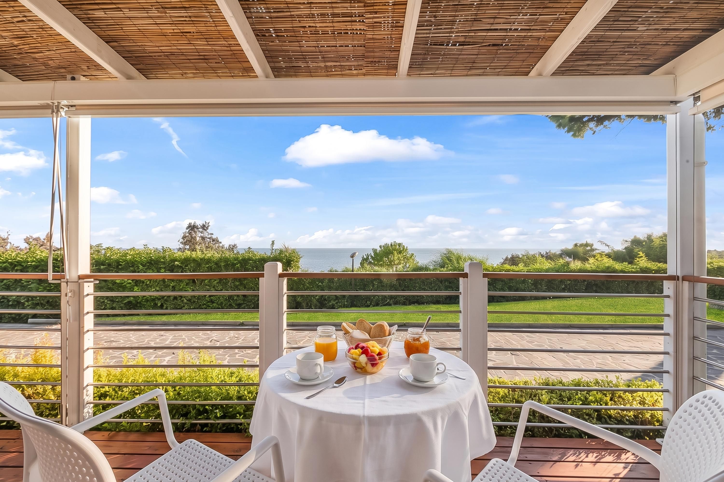 A table set for breakfast with a view of the sea, under a covered area with a bamboo ceiling.