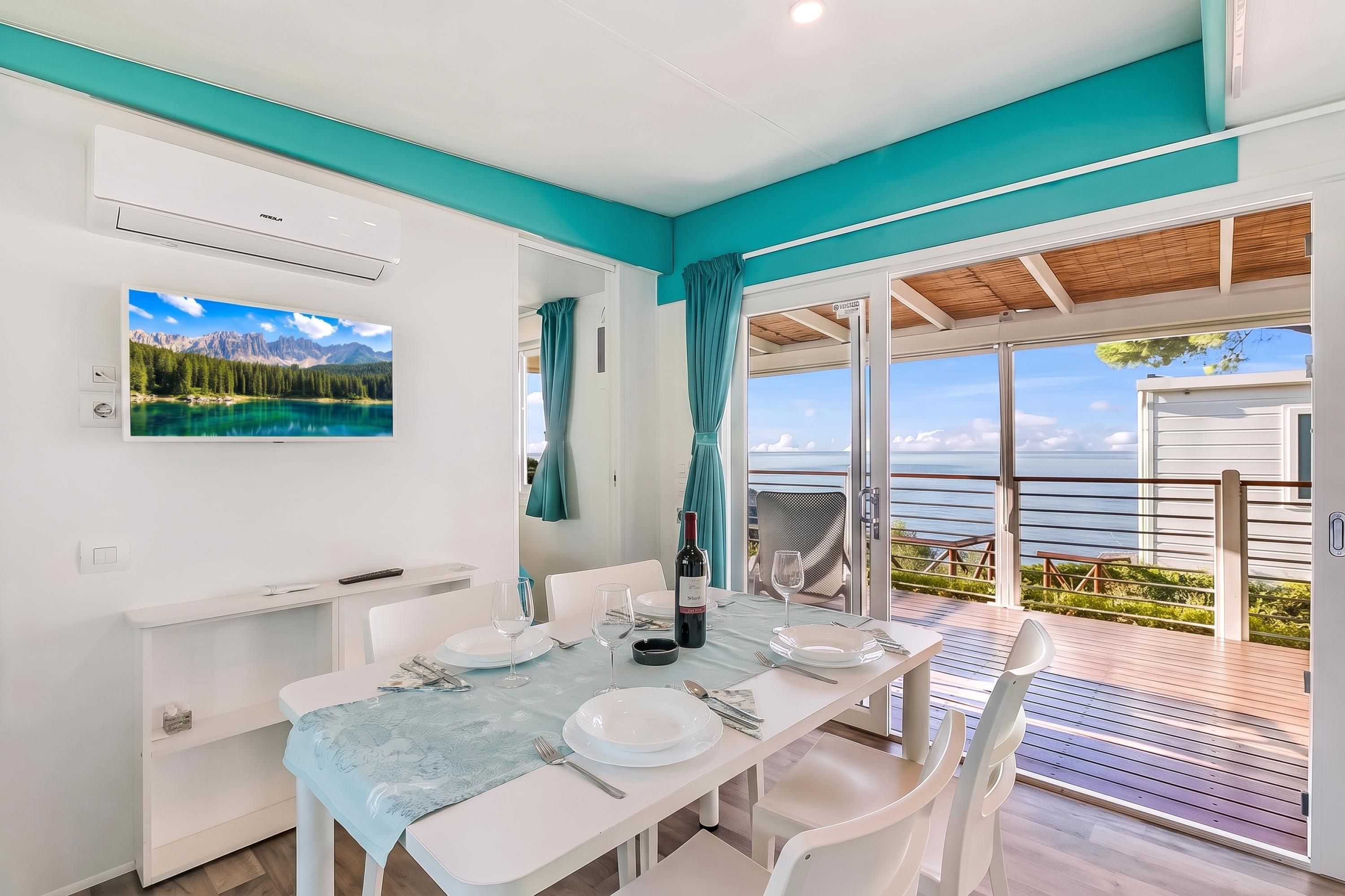 A dining area with a table set for two, a mounted TV, and a view of the sea through large glass doors.