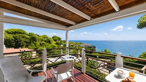 A balcony with a wooden ceiling, white chairs, a table with a croissant and juice, and a view of the sea and greenery.