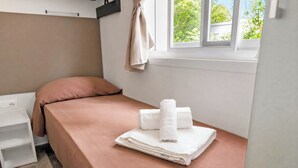 Un petit lit bien fait, avec une couverture rose et des serviettes blanches, une table de chevet blanche, un oreiller marron et une fenêtre donnant sur un paysage verdoyant.
