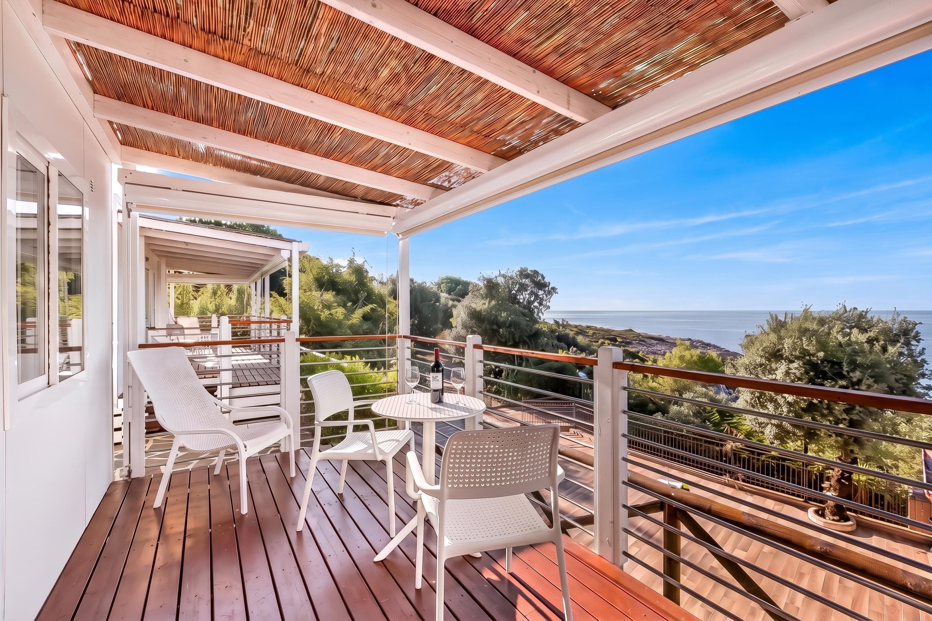 A covered outdoor area with a wooden deck, white chairs, and a table with a bottle and glasses.