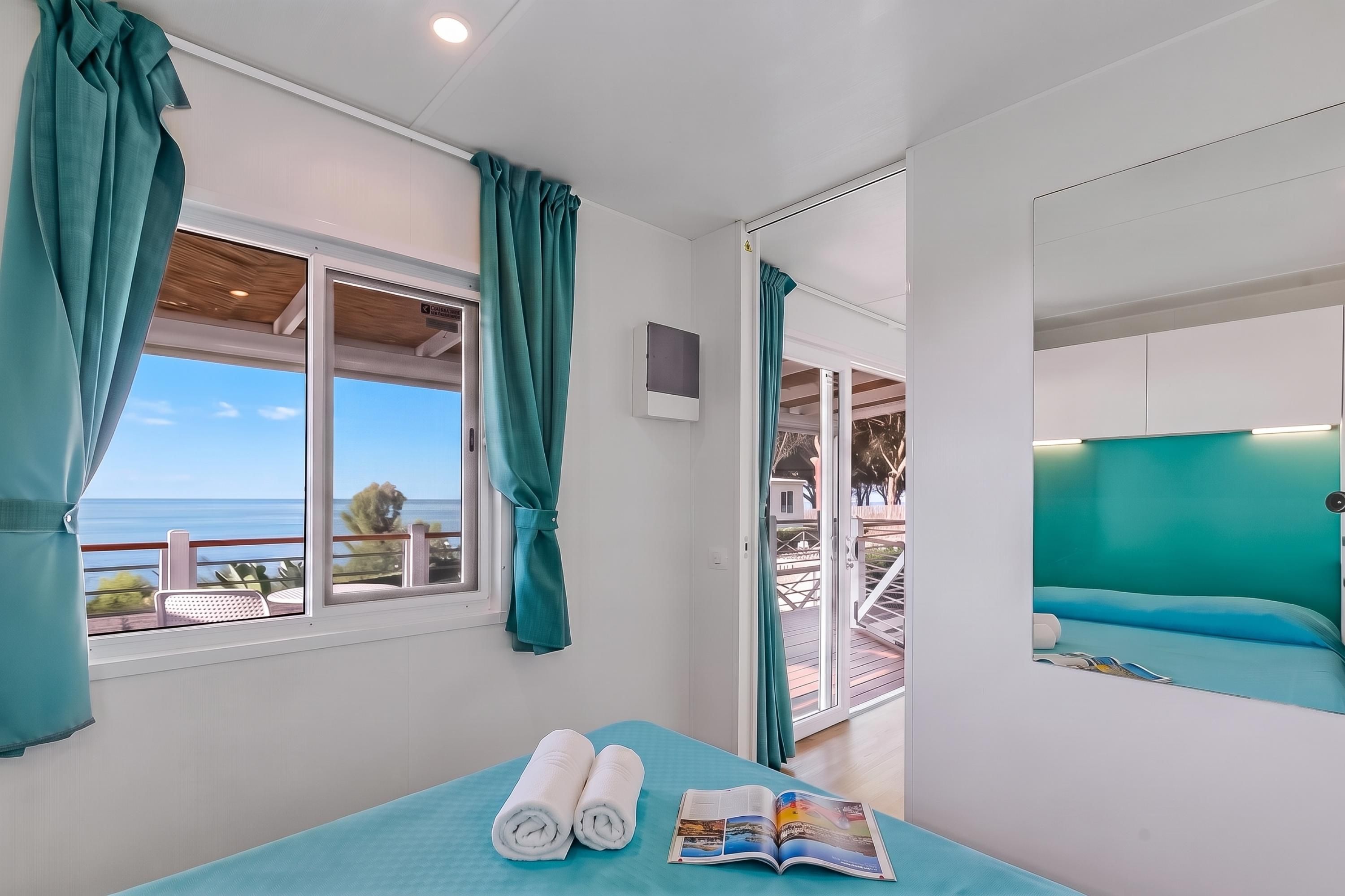 A room with a bed, a window with curtains, a view of the sea, and a magazine on the bed.