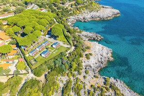 Vue aérienne d’une propriété située sur la côte, avec un rivage rocheux, un espace vert aménagé en parc et plusieurs bâtiments dotés de panneaux solaires.