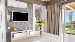 A modern room with a flat-screen TV mounted on the wall, a dining table with chairs, a vase with flowers, and a view of the outdoors through a door. - Sea View - 40 Parallelo - Deluxe 5, Marina di Camerota, Italy (Marina di Camerota)