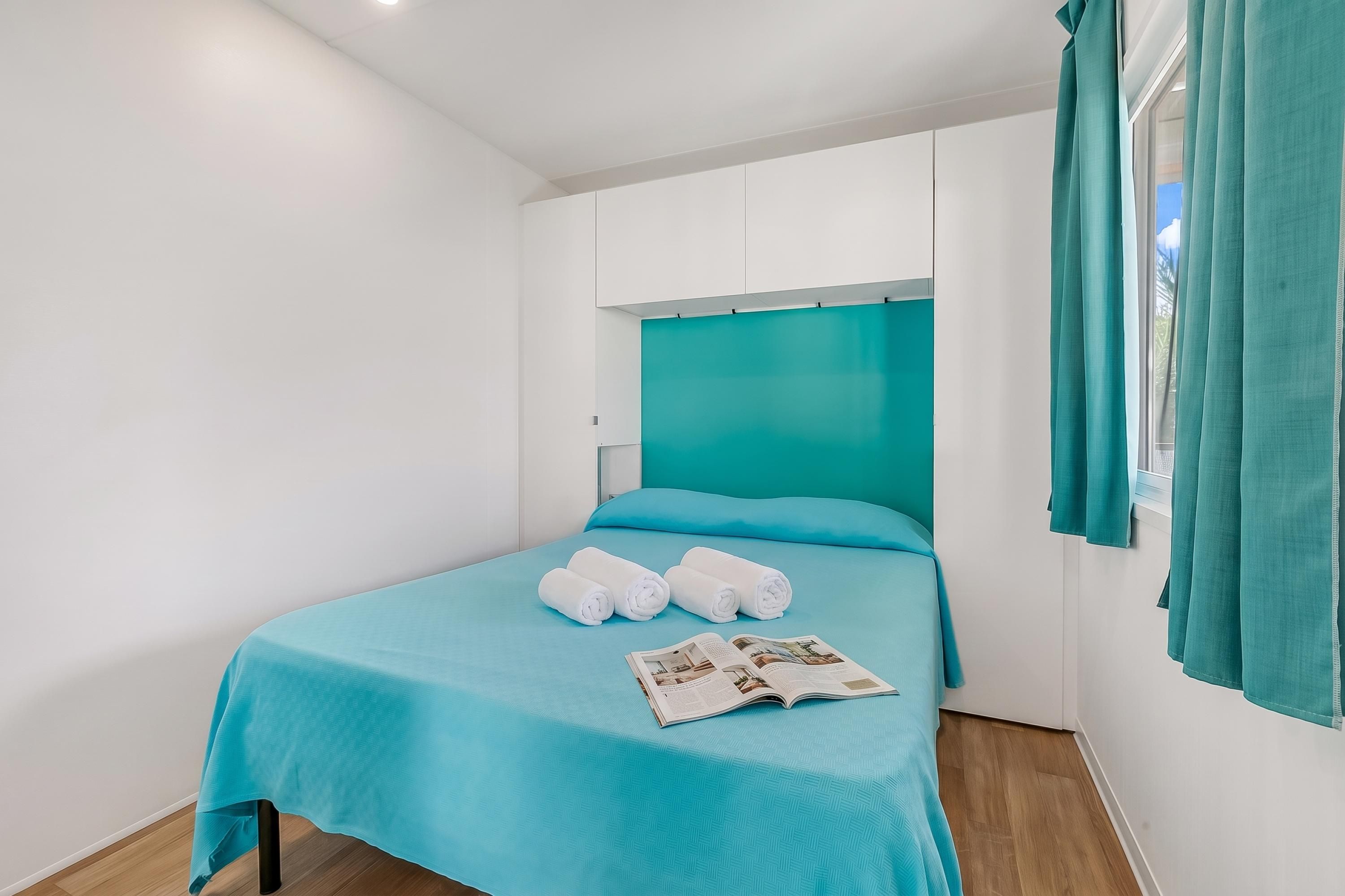 A single-bed room with a teal bedspread, white pillows, and rolled towels. A magazine is placed on the bed.