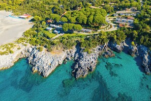 An aerial view of a coastal property with a beach, rocky shoreline, and clear turquoise waters.