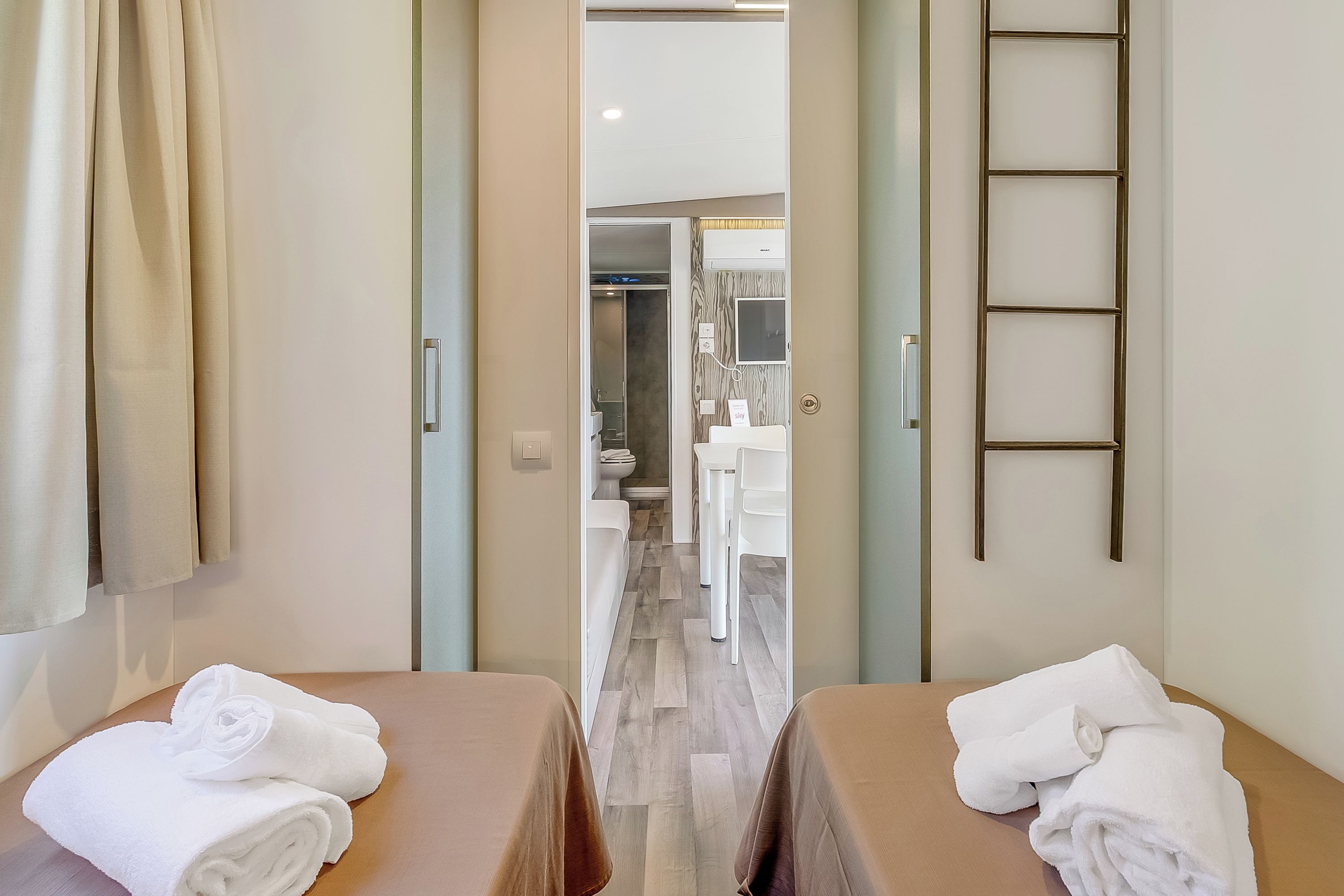 A hotel room with two beds, each with a towel, a bathroom with a mirror, and a shower area.
